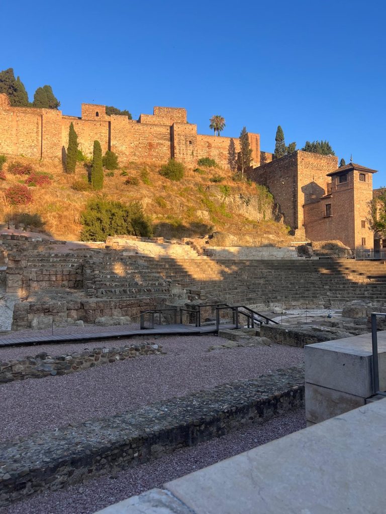 A vertical shot of the Roman Theatre in Malaga, Spain, with the Alcazaba fortress in the background. The Roman Theatre's tiered stone seats and ruins are in the foreground, with a walkway crossing the center. The Alcazaba is a large, medieval brick fortress situated on a steep hill above the theater. The warm, golden light of the late afternoon sun illuminates the brickwork of the fortress and casts long shadows across the ruins below. The sky is a clear, deep blue.
