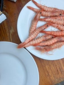 A wide-angle, top-down shot of a large plate of cooked shrimp with their heads and tails still on, served on a rustic wooden table. The pinkish-orange shrimp are scattered on a white plate and sprinkled with coarse sea salt. An empty white plate is visible in the bottom left corner.