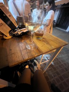 Cheila´s point-of-view shot from a table on a narrow cobblestone street at night. A large wine glass with a small amount of white wine and a shorter glass with a dark liquid are on a rustic wooden table. Below the table, Cheila´s legs and Julião, is visible. Two people are standing in the background behind the table, a man with a mustache and a woman with long, dark hair, both looking toward each other. A menu on a stand is on the left side of the table.