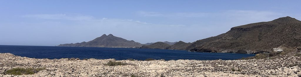 Panoramic view of an arid coastal landscape, with deep blue sea in the foreground and brown rocky mountains in the background under a clear blue sky.