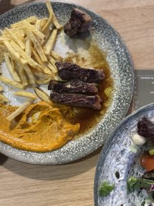 A close-up, top-down shot of a partially eaten plate of food. The main plate, a gray-blue ceramic dish, holds a few pieces of sliced, medium-rare steak covered in a glossy brown sauce. To the left are a pile of golden French fries and a creamy, orange-colored sauce. The plate is smeared with sauces and grease. In the bottom right corner, a portion of another plate containing a salad is visible.