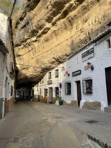 A street-level shot in Setenil de las Bodegas, Spain, showing the unique architecture of the town. A narrow, paved street is flanked on the right by a row of traditional whitewashed buildings with brown wooden doors and black-barred windows. The buildings are nestled directly under a massive, overhanging rock cliff that serves as their roof, creating a "cave street" effect. The golden afternoon sunlight illuminates the underside of the rock formation, highlighting its textured surface. The street is empty of people, and signs for a restaurant called "La Cuevecita" are visible on the buildings