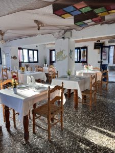 A warm, sunlit interior shot of a casual seafood restaurant. The dining room has a white ceiling with a decorative fishing net draped across it and exposed light bulbs hanging down. The walls are also white, one featuring a painted mural of an octopus and a shark. Several wooden tables, each covered with a white tablecloth, are set for dining with plates, glasses, and small floral arrangements. The floor is made of mottled gray and brown tiles. Sunlight streams in from a window on the left, illuminating the space. There is a small counter or bar area in the background with a person visible.