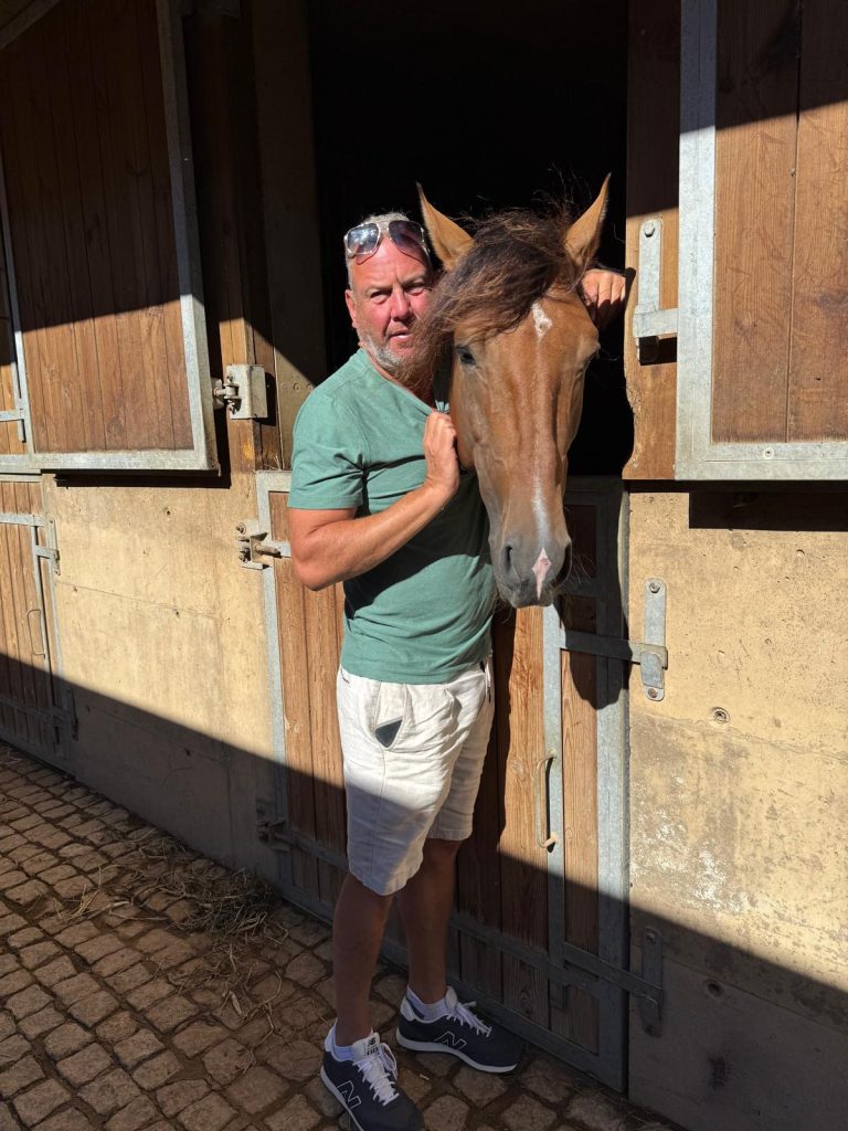 Alan stands in the doorway of a wooden stable, embracing a horse's head that is leaning out of the stall. Alan is wearing a green t-shirt, white shorts, and has sunglasses resting on his head. The photo is taken in bright sunlight, highlighting the warm tones of the horse's chestnut coat and mane, and the wooden stable door.