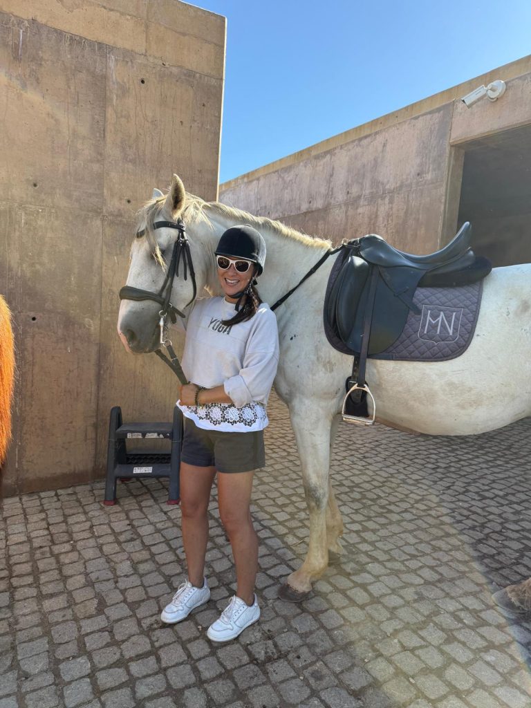Cathy is pictured outdoors, standing next to a white/light grey horse, ready for riding. She's wearing a black riding helmet and sunglasses, a light-colored long-sleeve sweatshirt with a lace-trimmed hem, olive green shorts, and white sneakers. The horse is saddled with a black saddle and a dark pad featuring a white monogram. They are standing on a cobblestone ground with a textured, sunny-colored wall in the background. It looks like a bright day at the stables!