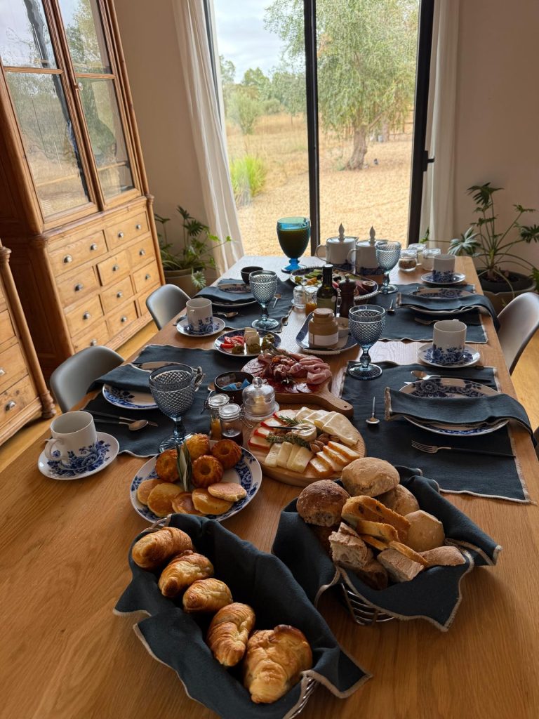 A delightful and well-presented breakfast or brunch spread on a wooden table. A central wooden paddle board is laden with an assortment of sliced cheeses, garnished with nuts and rosemary. To the right, another board holds various cured meats. On the left, a plate features golden-brown pastries and pancakes.