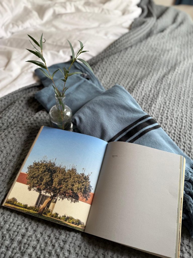 The main subject is an open book resting on a grey knit blanket. The left page displays a photograph of a white house with a terracotta roof and a large tree against a blue sky, while the right page is a blank grey swatch, possibly labeled with a Pantone color (like 7544). Next to the book is a small glass vase holding a few green sprigs. Behind the arrangement, a dusty blue blanket with dark stripes is folded, set against a background of white sheets.