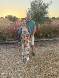 Cathy and Alan smiling and standing on a gravel path at sunset, surrounded by wildflowers and olive trees at Malhadinha Nova in the Alentejo countryside.