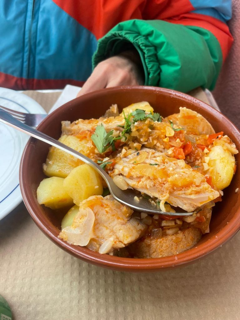 A rustic bowl of ensopado de raia, with tender pieces of ray in a rich, reddish broth, served with boiled potatoes, bread, onions, garlic, peppers, and fresh coriander on top. A spoon rests inside the bowl, with a person in a colourful jacket seated behind it