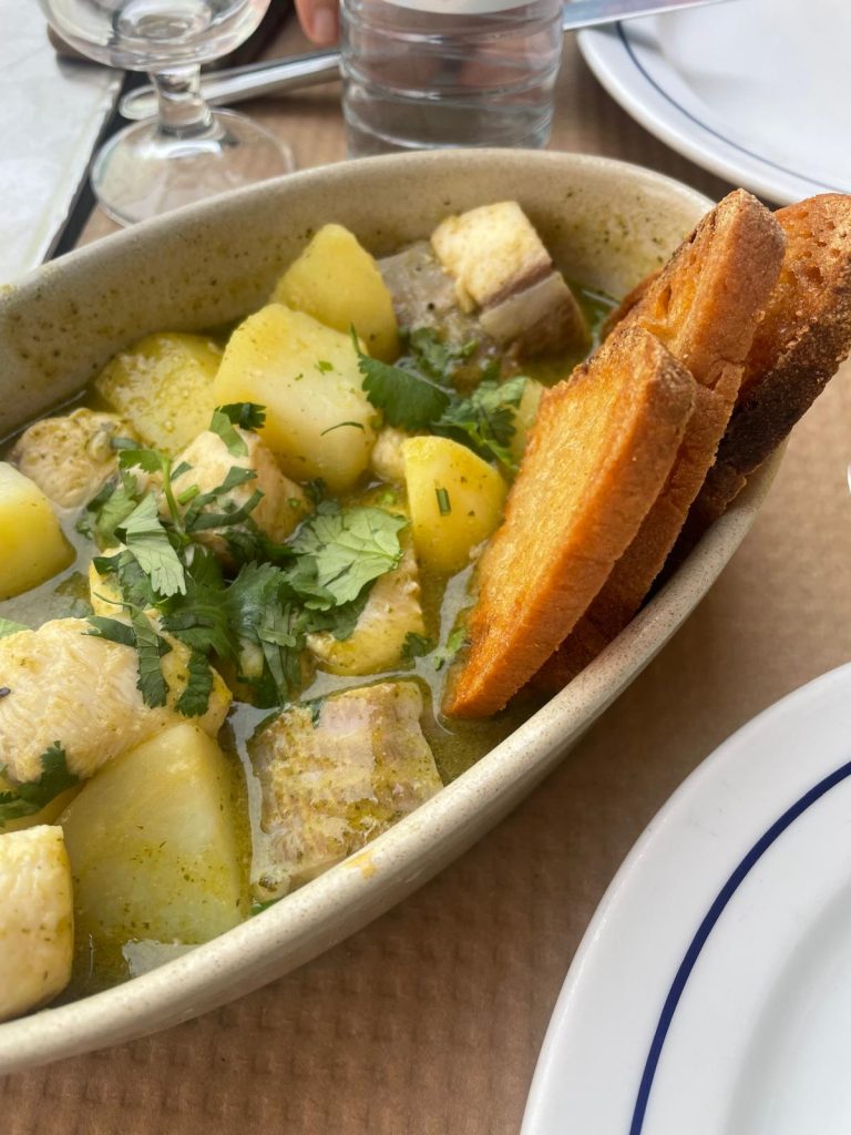 A bowl of coentrada de cação, with chunks of tender dogfish in a light, aromatic coriander broth, served with boiled potatoes and slices of toasted bread resting on the edge of the bowl. Fresh herbs are scattered on top, with a simple table setting in the background