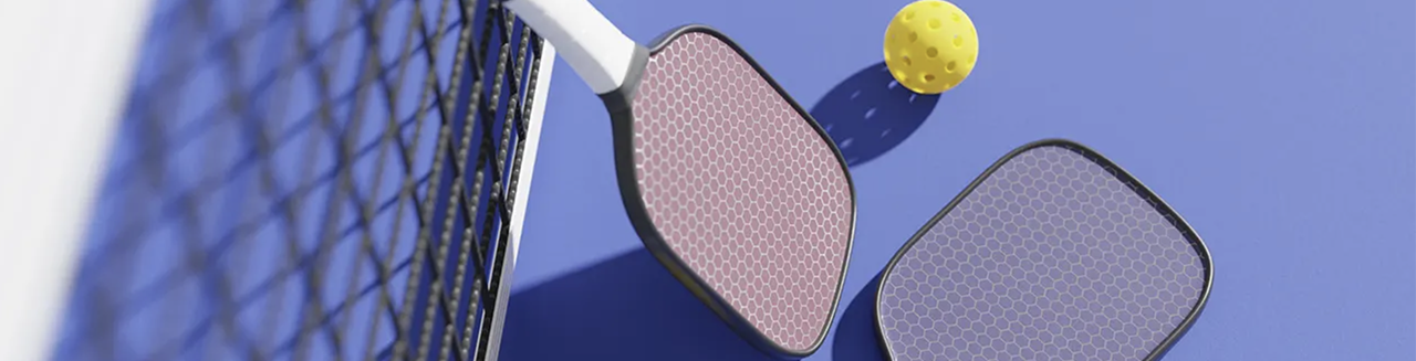 Two pickleball paddles — one pink and one purple — lie on a blue court beside a net, with three yellow pickleballs scattered around.